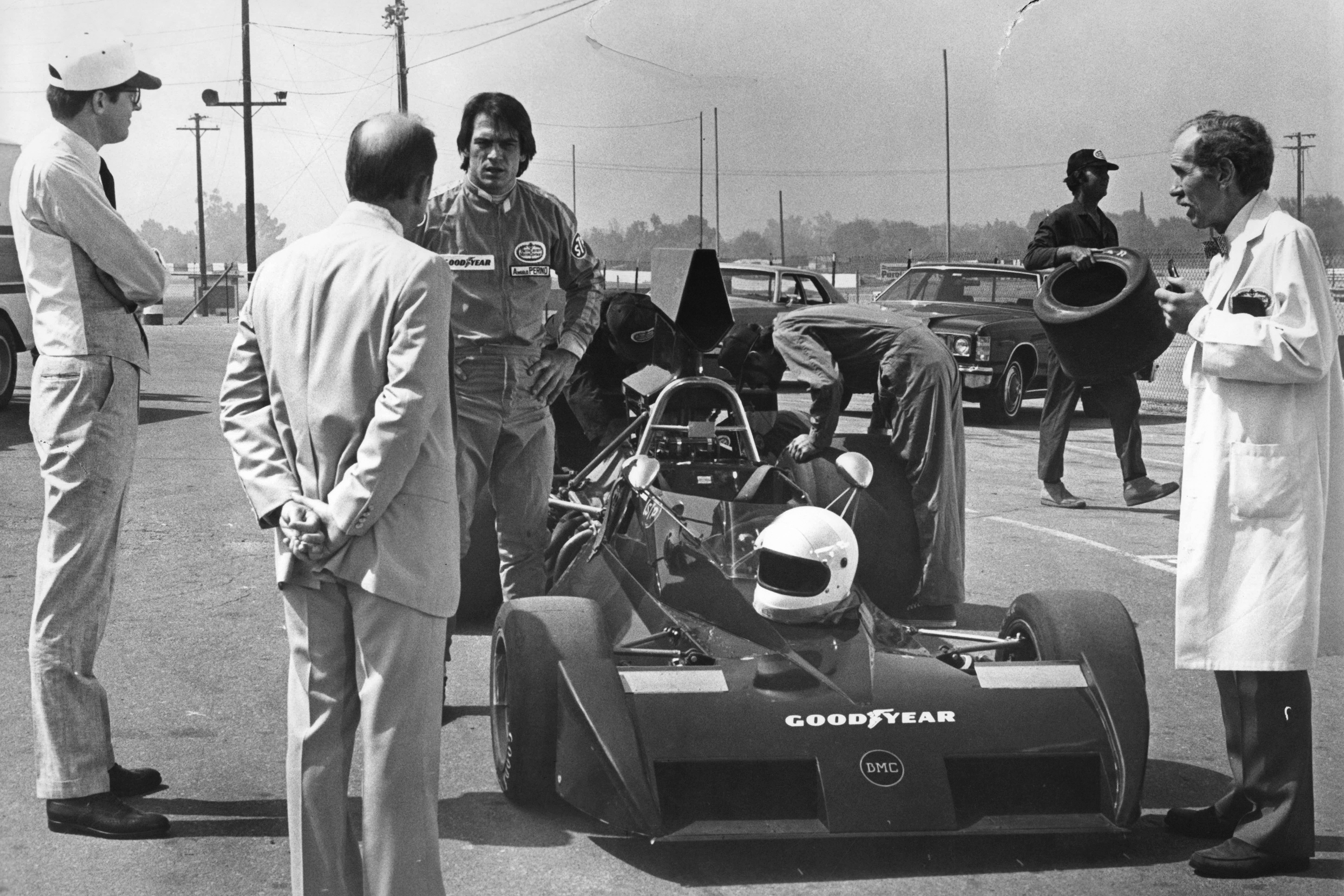 Vintage race car in the pit surrounded by crew members inspecting it, Goodyear logos visible on the car and drivers in period uniforms.