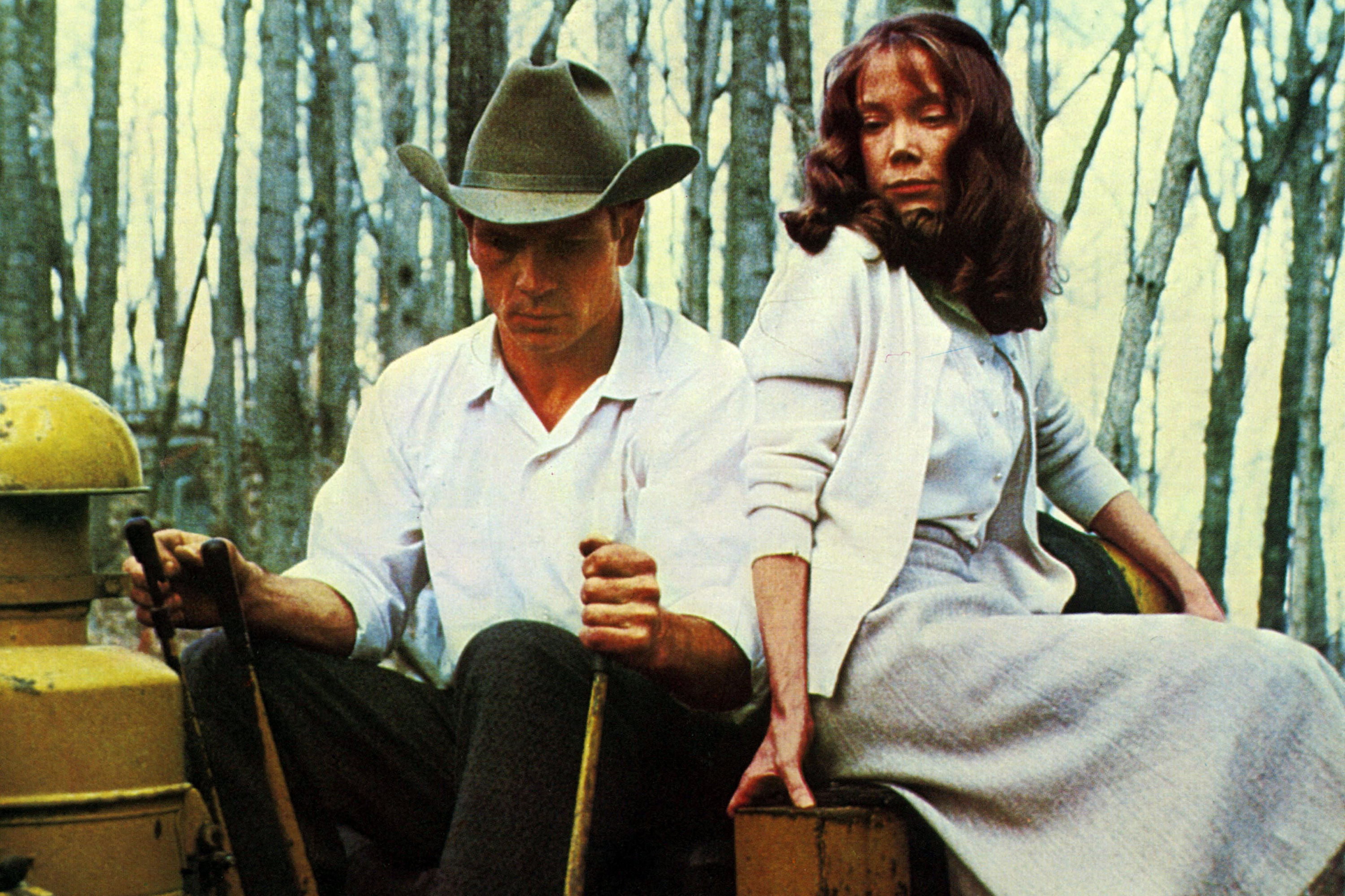 Man and woman in a rowboat paddling through a forest of bare trees; man in a hat and white shirt, woman with long hair beside him.