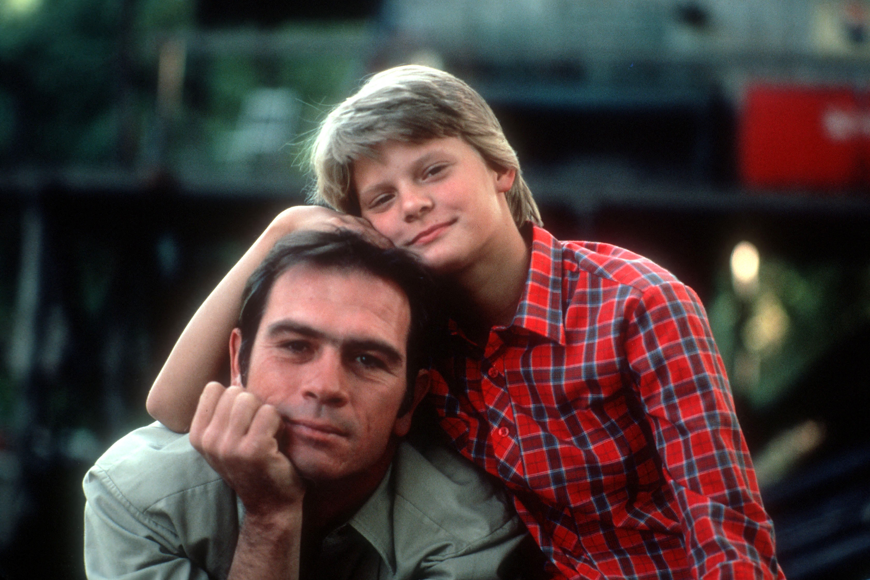 Boy in a red plaid shirt rests his head on a man's shoulder as they share a hug and smile at the camera.