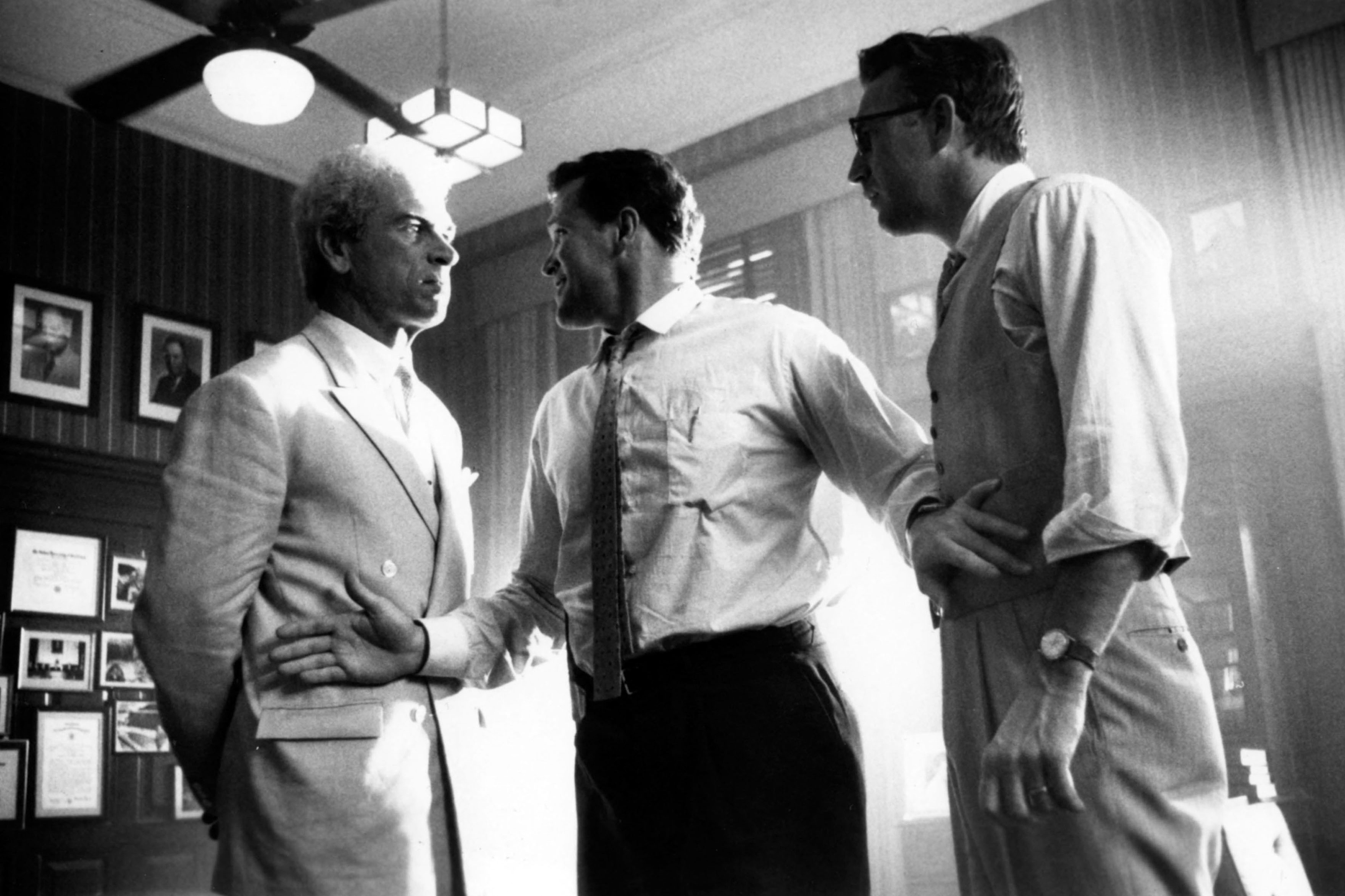 Three men in business attire shake hands in a wood-paneled office, framed photos on the wall behind them.