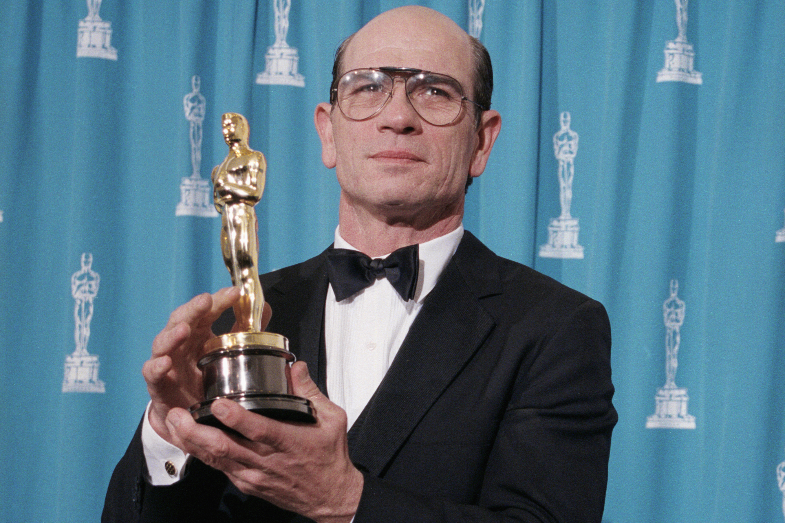 Man in a tuxedo holding an Oscar statuette, with a turquoise backdrop featuring award logos, facing camera.