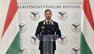 Uniformed official at a podium during a press conference, Hungarian flags on both sides and a backdrop reading 'ALKOTMÁNYVÉDELMI HIVATAL'.