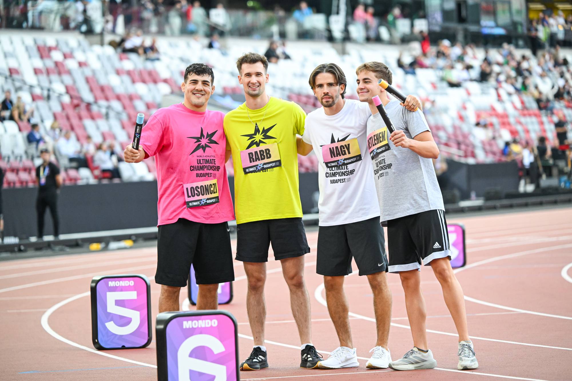 Four male athletes stand arm-in-arm on a running track, each holding a relay baton and smiling at the camera during a race event. Their colorful shirts and bib numbers are visible.