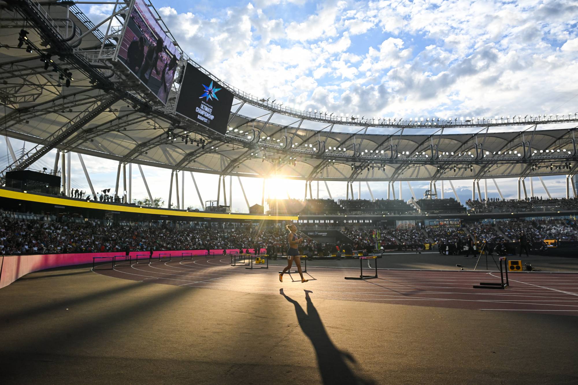 Athlete walking on a curved running track inside a large stadium with a digital screen and audience in the stands, sunset lighting.