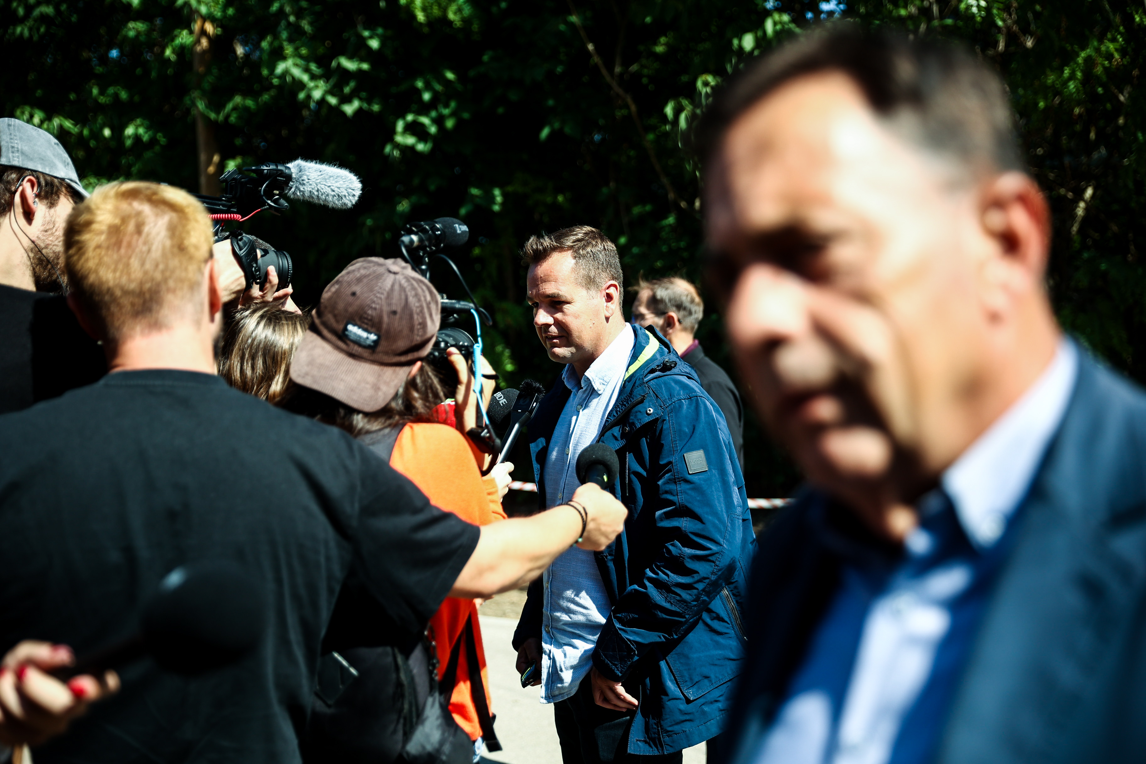 Man being interviewed by multiple reporters as cameras and microphones surround him outdoors.