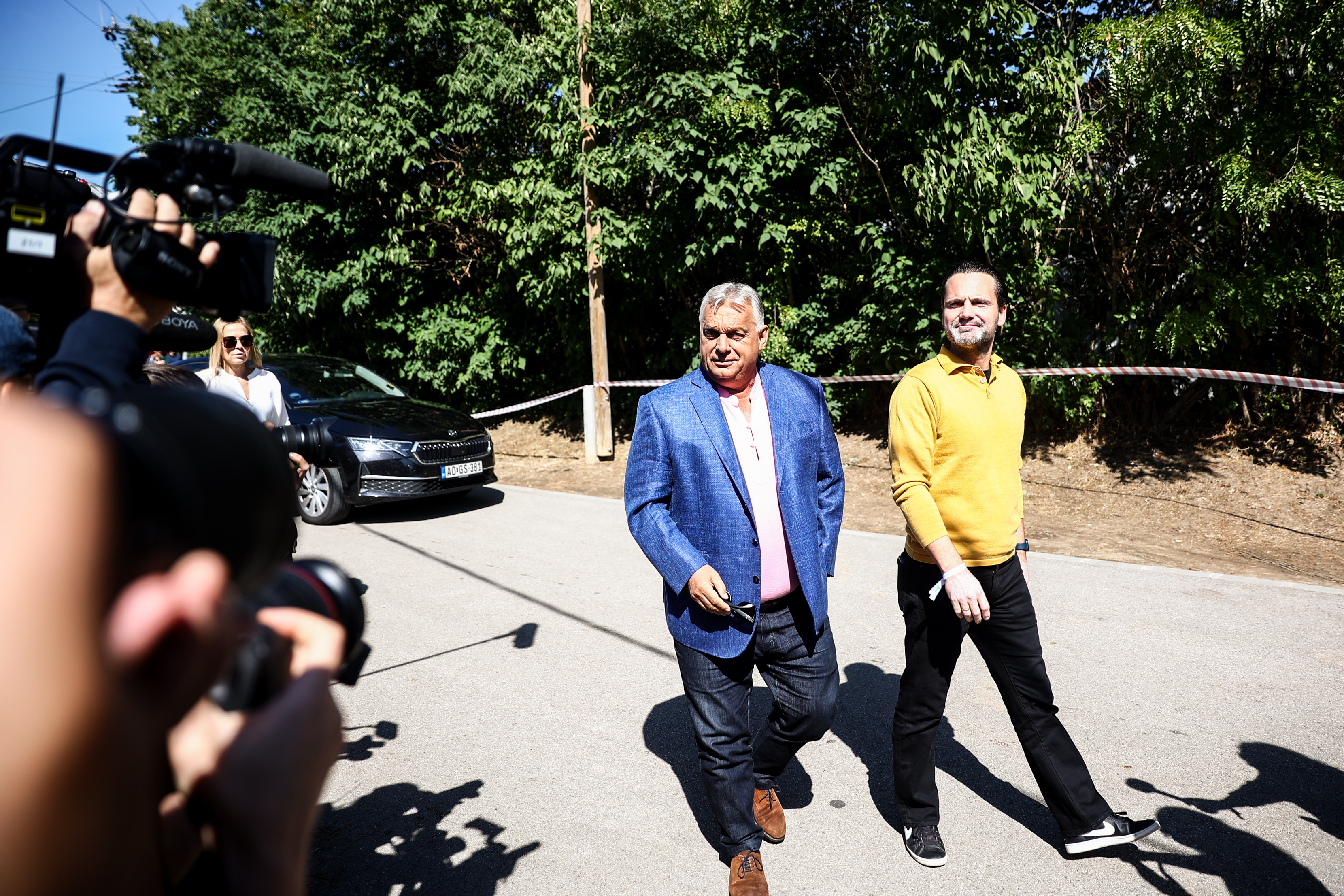 Two well-dressed men walk outside as photographers with a video camera crowd them and film the scene under a sunny sky.