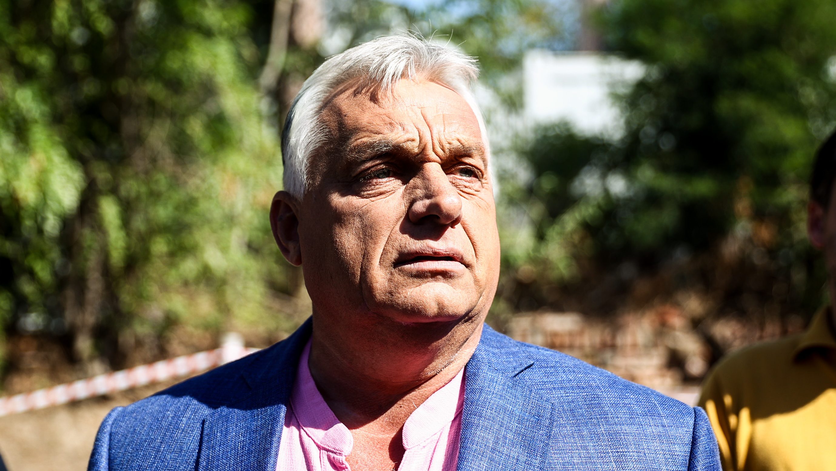 Close-up of an older man with white hair, wearing a pink shirt and blue blazer outdoors.