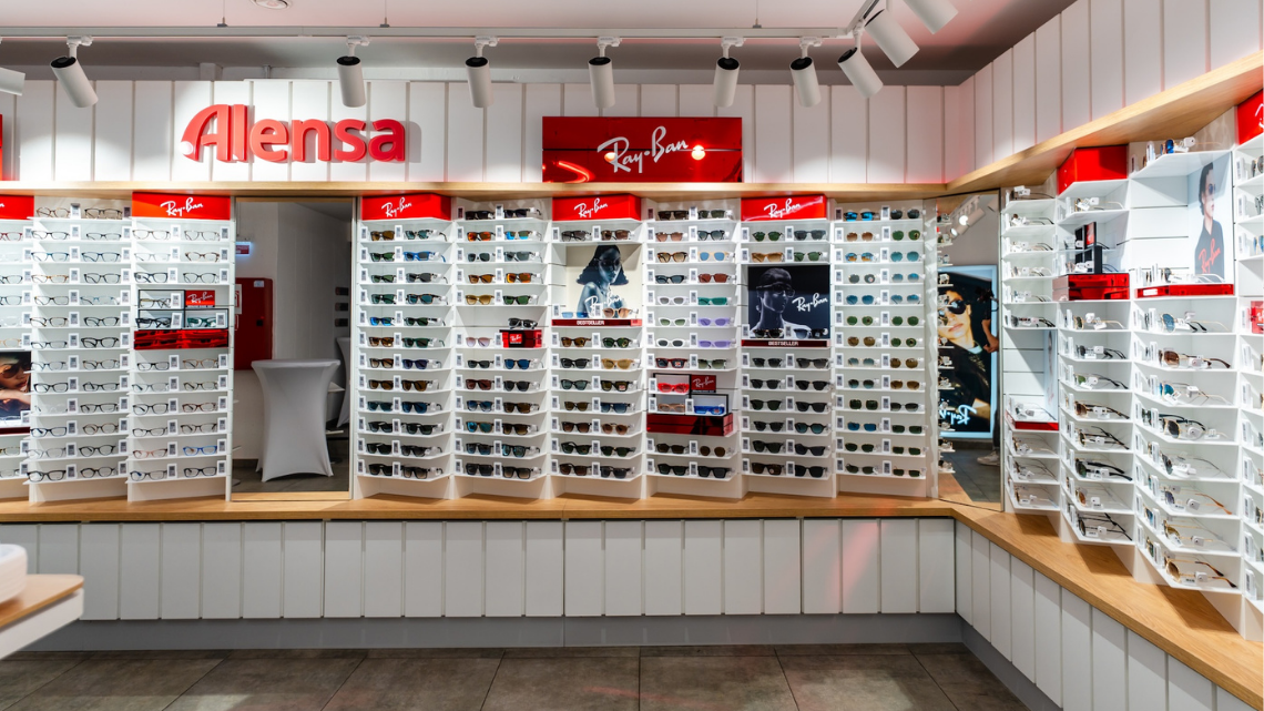 Interior of a bright sunglasses store with white display racks filled with Ray-Ban and other sunglasses, a wooden counter, and brand signage above.