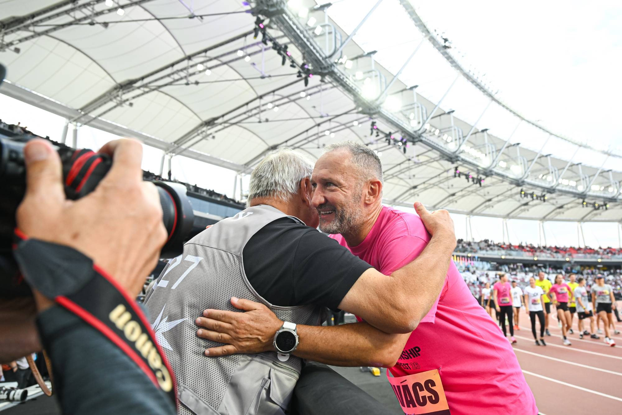 Two men hugging on a track at a sports event, with a photographer in the foreground capturing the moment
