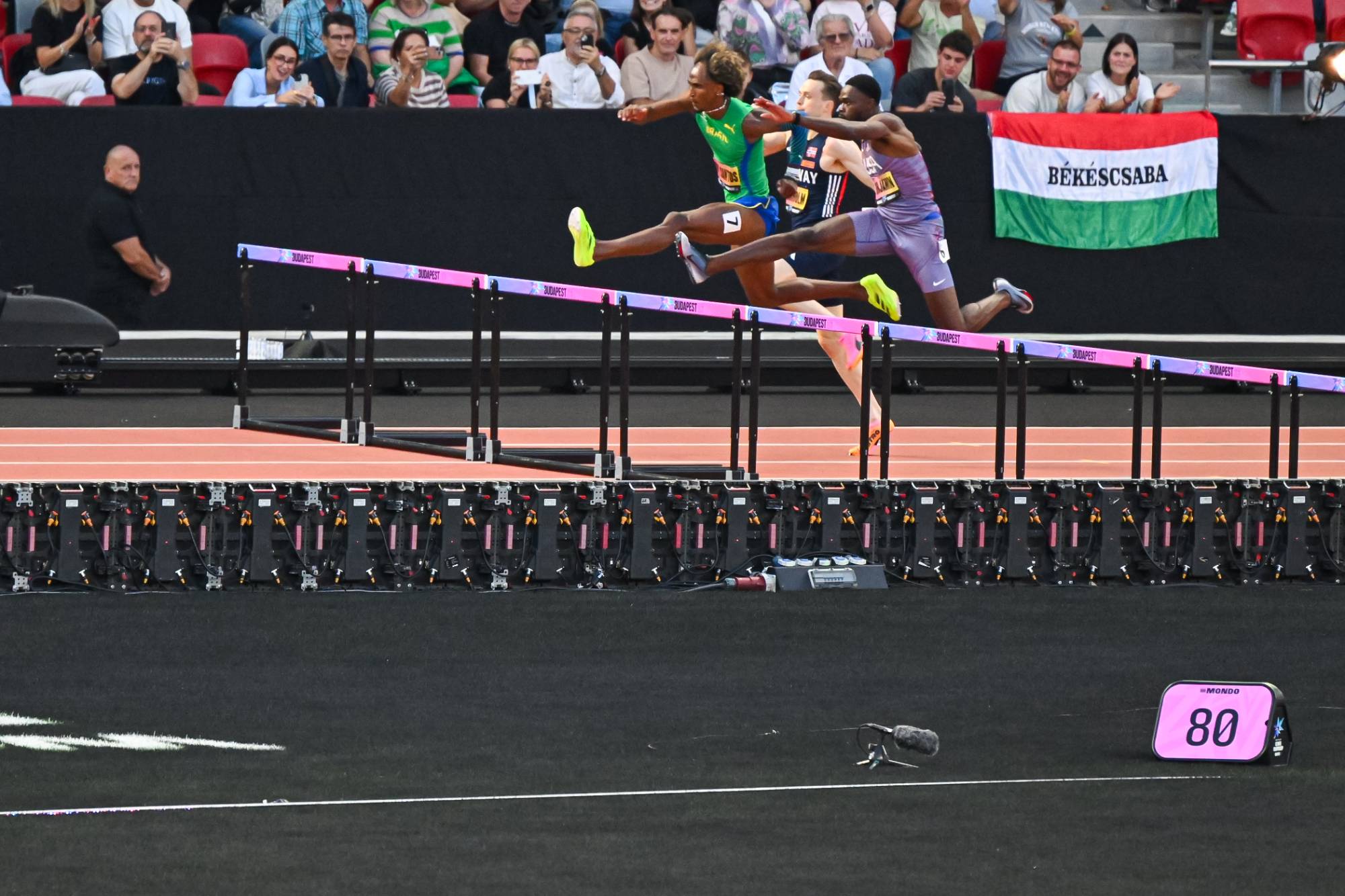 Three male athletes leap over hurdles in a sprint race as a crowd watches from the stands, with a Békéscsaba flag visible in the background.