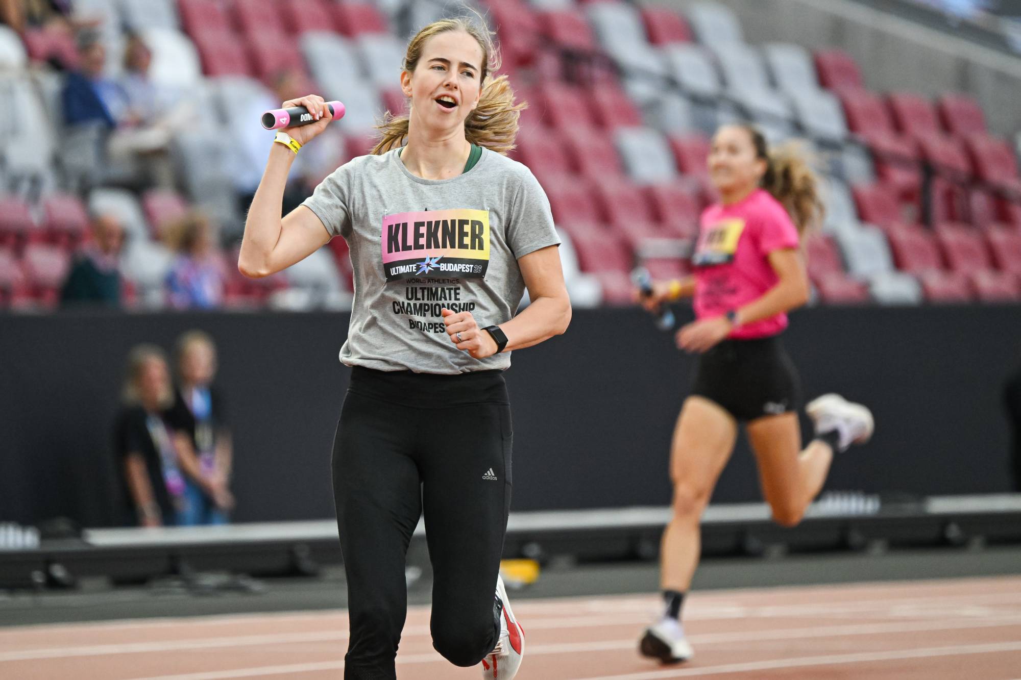 Female runner in a gray shirt with 'KLEKNER' visible, sprinting on an indoor track with blurred spectators in the background.