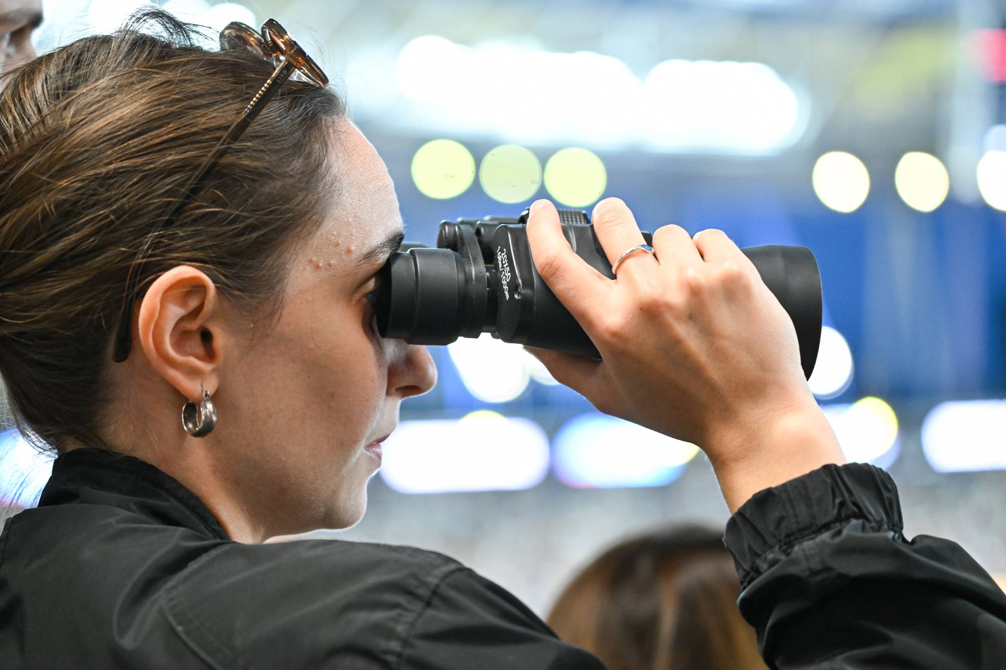 Woman using black binoculars up to her eye at an indoor event, sunglasses resting on her head.