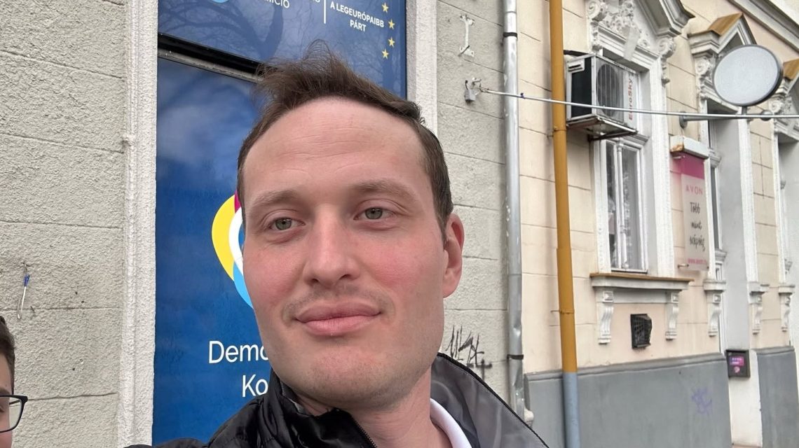 Close-up of a man taking a selfie on a city sidewalk, with a blue storefront poster behind him and an ornate building facade.