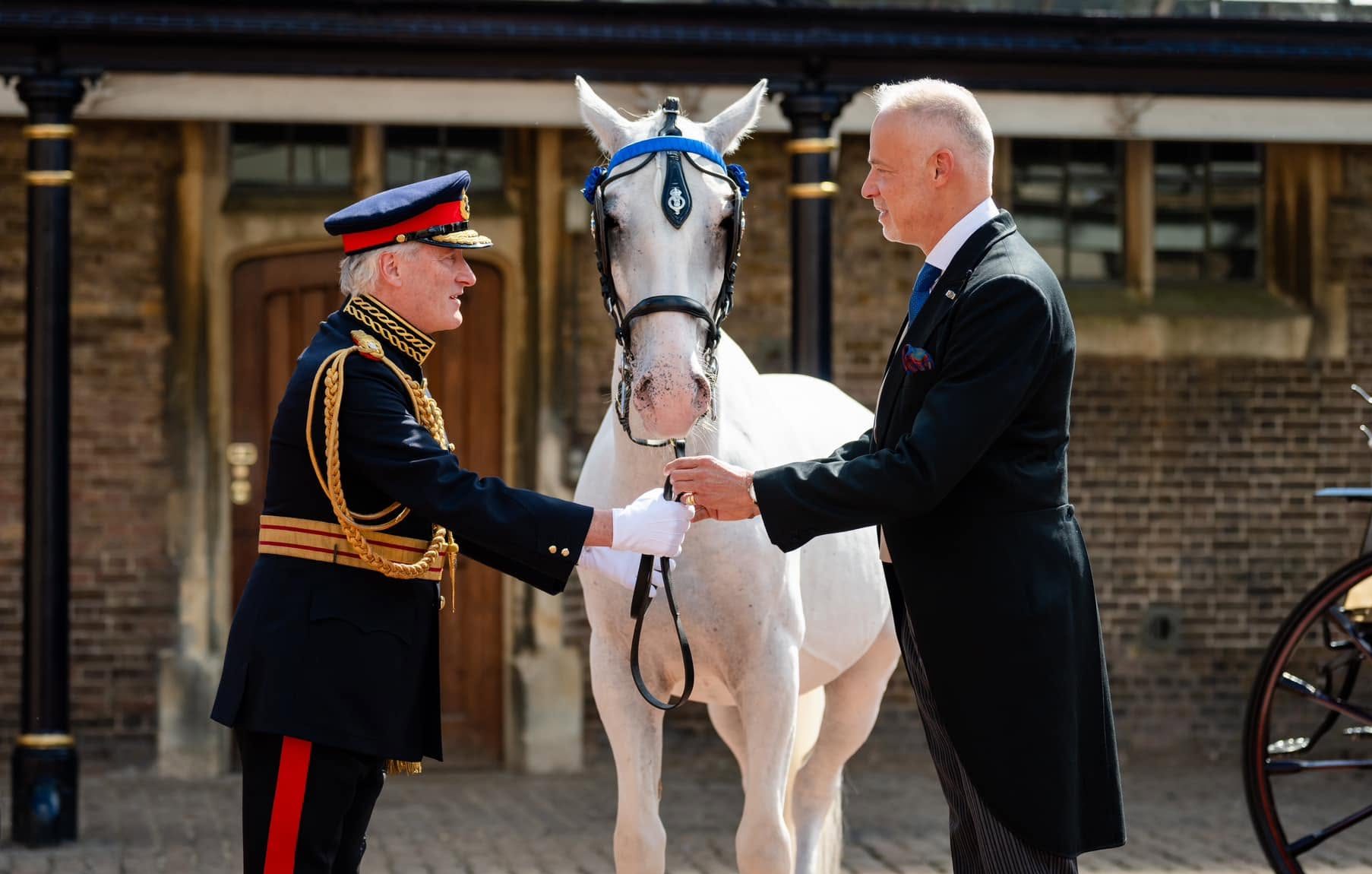Two men in formal dress shake hands beside a white horse outside a brick building