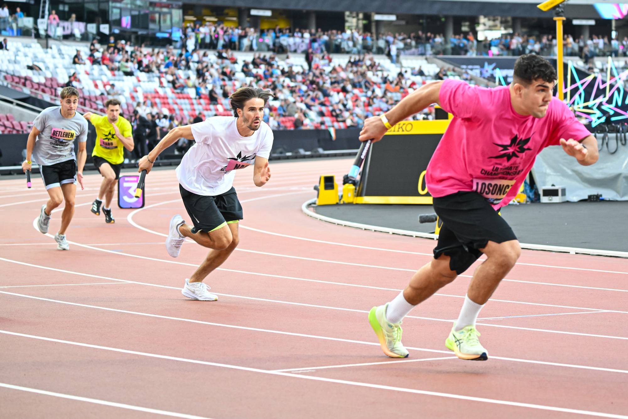 Four male sprinters race on a red track with a cheering crowd in the stadium stands behind them.