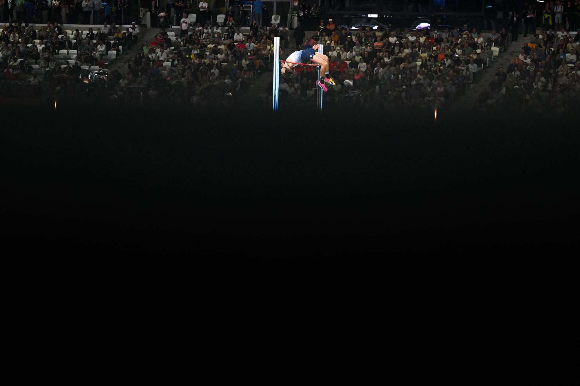 Gymnast mid-air on a horizontal bar during a gymnastics routine, with a large audience watching in an arena.