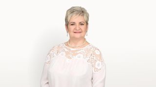 Smiling middle-aged woman with short gray hair, wearing a white lace blouse and pearl necklace against a light background.