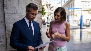 Man in a navy suit and tie reviews papers with a woman in a pink sleeveless top on a city sidewalk.