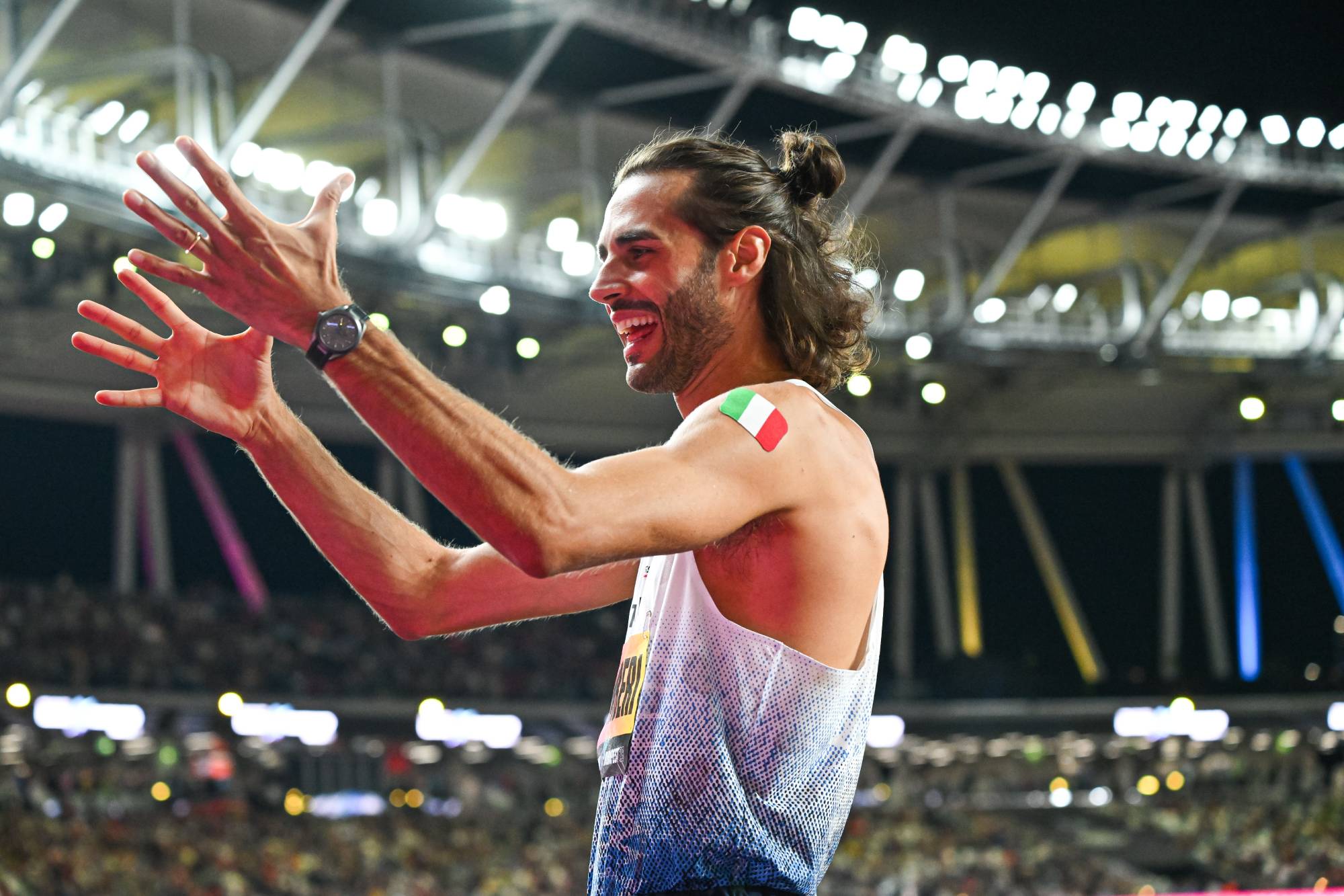 Athlete with long hair tied back celebrates with arms raised in a stadium, Italian flag on shoulder.