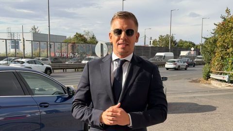Man in a dark suit and sunglasses stands beside a blue car on a busy street, posing for the camera.