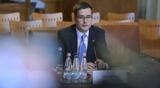 Man in a suit and glasses speaks into a microphone at a conference table with bottled water and a name plate in front of him