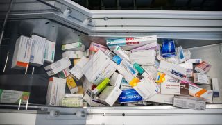 Top-down view of a metal medicine cabinet overflowing with scattered pharmaceutical boxes in a drawer, mixed brands and dosages.