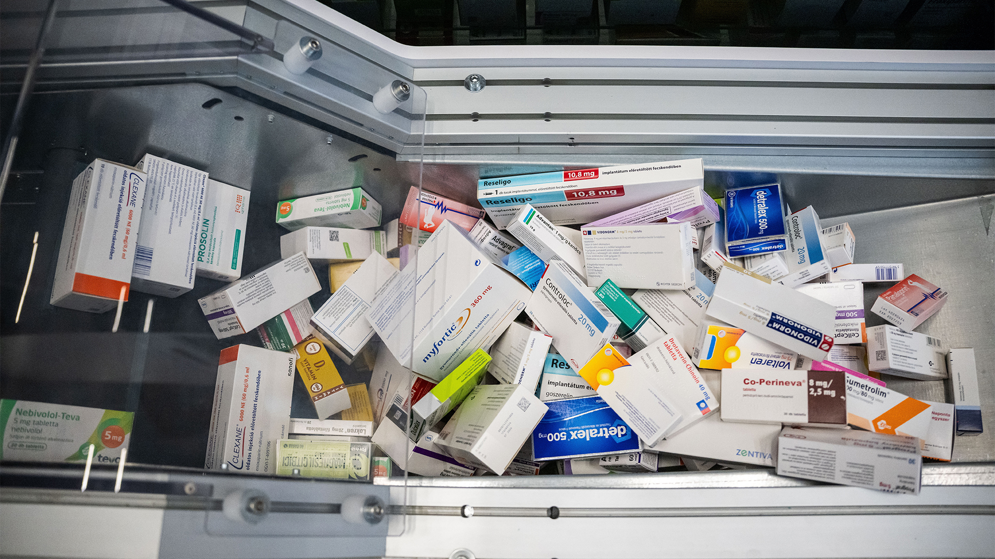 Top-down view of a metal medicine cabinet overflowing with scattered pharmaceutical boxes in a drawer, mixed brands and dosages.