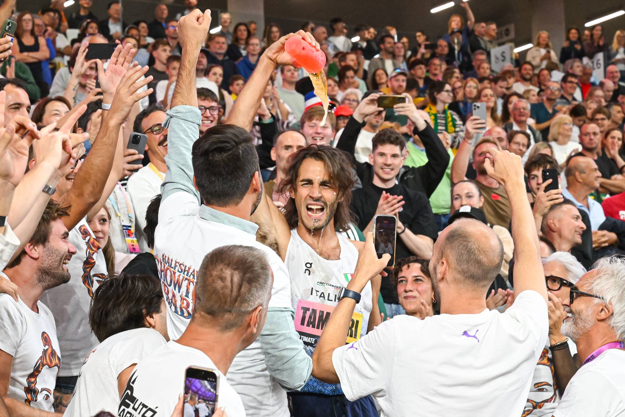 A man in a white shirt pours liquid from a pink bottle toward a cheering crowd at a sporting event.