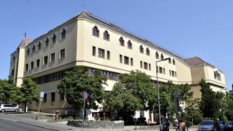 Large beige multi-story building with arched windows, trees in front, and cars parked along a sunny street.