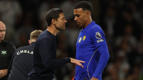 Two men face off on a soccer field, one in a blue Chelsea jersey and the other in dark clothing, leaning in with tense expressions as their hands meet.