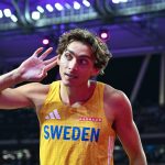 Swedish sprinter in a yellow jersey with 'SWEDEN' on the chest, cupping ear while competing under bright stadium lights.