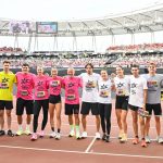 Group of runners in bright yellow, pink, white, and gray shirts pose arm-in-arm on a stadium track, with a large crowd in the stands behind them.