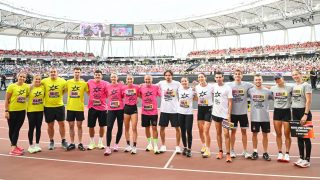 Group of runners in bright yellow, pink, white, and gray shirts pose arm-in-arm on a stadium track, with a large crowd in the stands behind them.