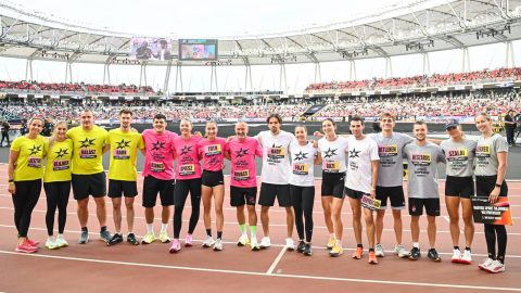 Group of runners in bright yellow, pink, white, and gray shirts pose arm-in-arm on a stadium track, with a large crowd in the stands behind them.