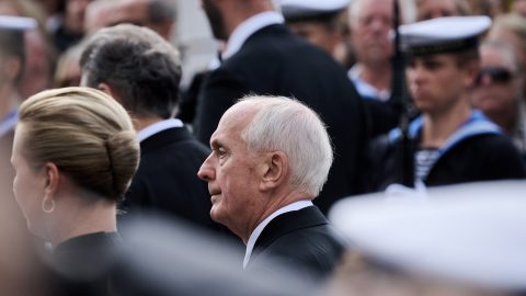 Side profile of an older man with white hair in a suit at a formal crowd event, with uniformed personnel blurred in the background.