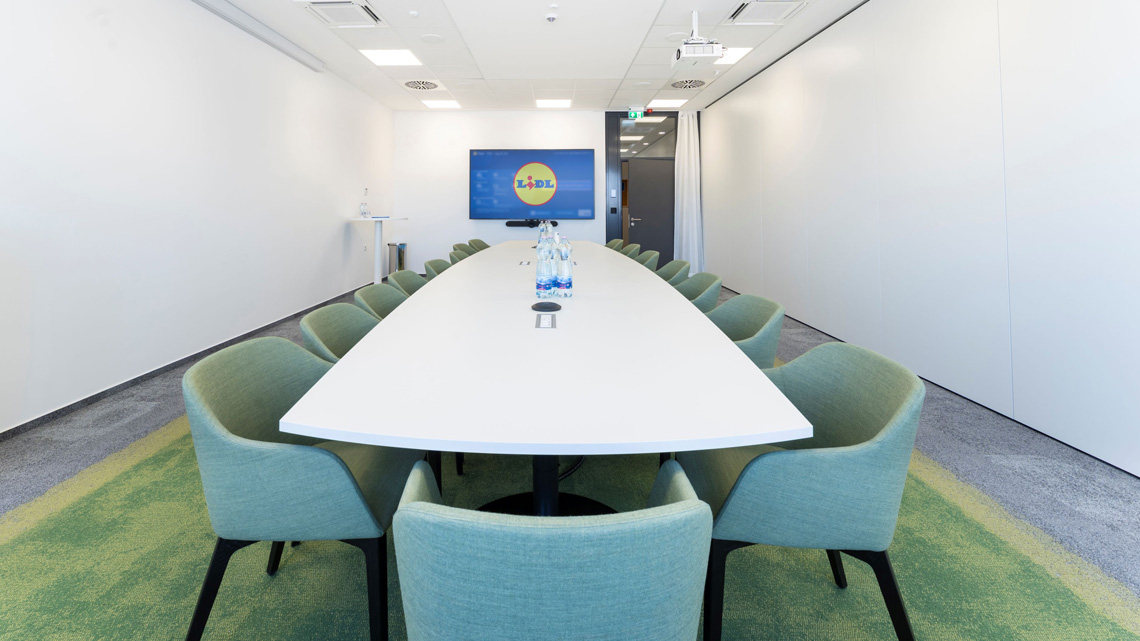 Modern meeting room with a long white oval table, green upholstered chairs, and water bottles, a projector screen showing Lidl in the background.