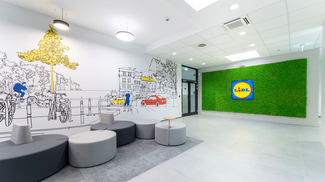 Lobby area with a green moss wall and Lidl display, circular gray seating, and a line-drawing city mural on the wall.