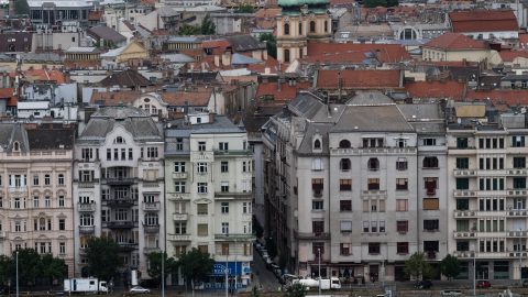 Dense European cityscape with red-tiled roofs, ornate facades, and a green-domed church tower in the distance