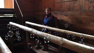 Woman in a blue jacket and red backpack sits on a wooden railing inside a rustic log cabin, with bubbles floating around.