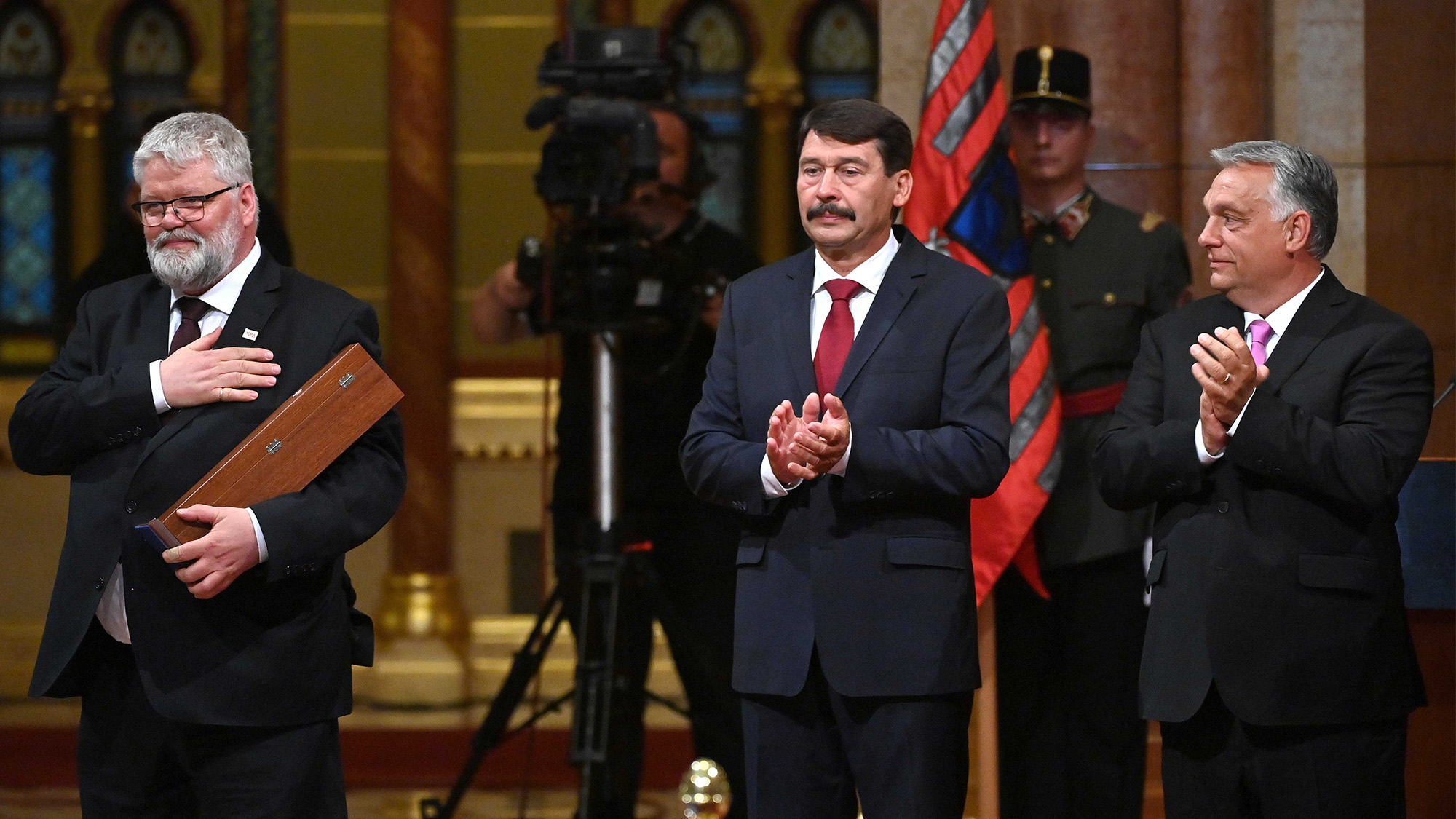 Three men in suits at a formal ceremony, with one holding a wooden award plaque while the others applaud; US flag in the background.