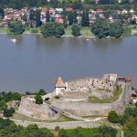 Castle ruins on a hill overlooking a wide river, with a town along the far shore and several boats on the water.