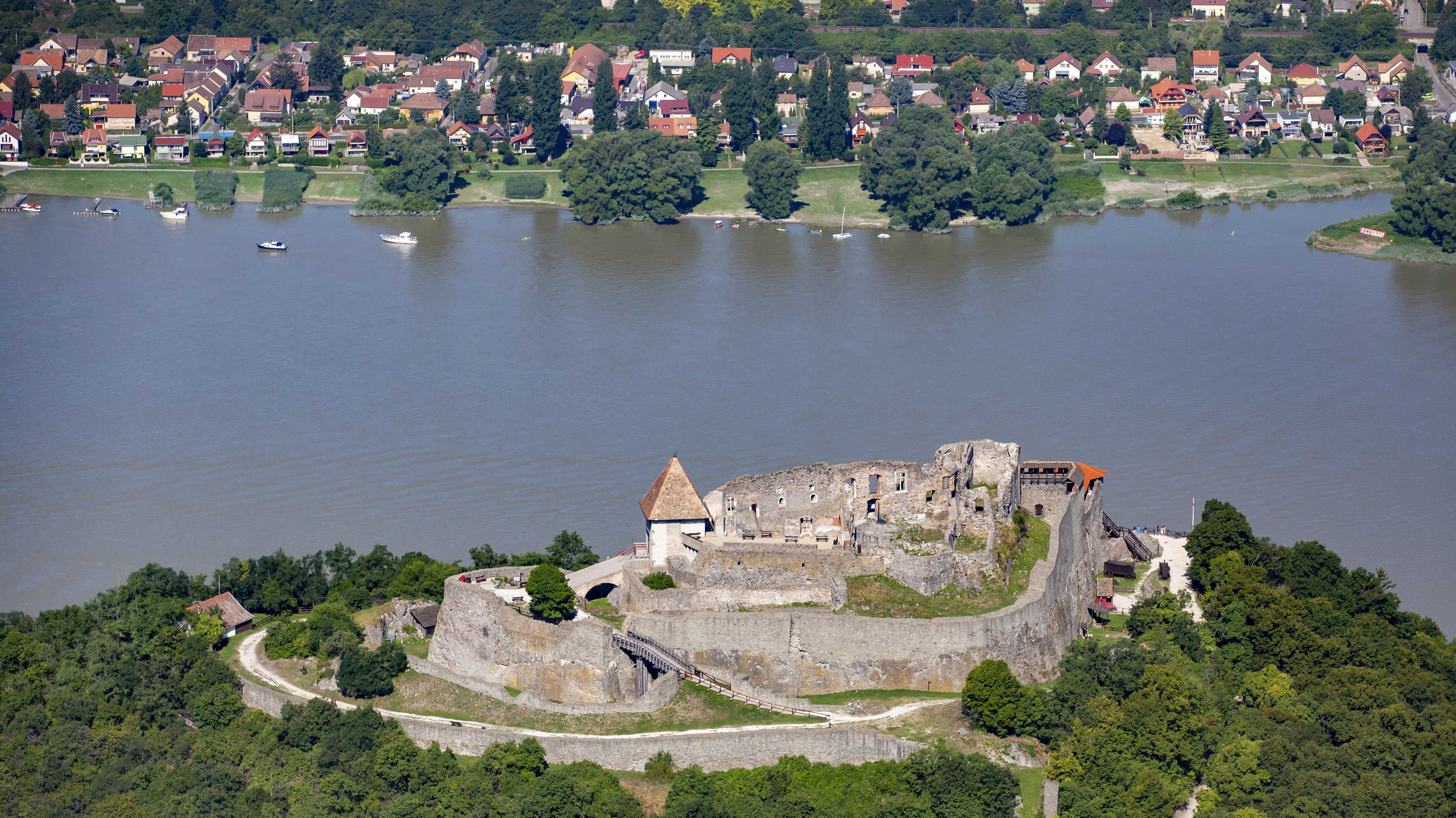 Castle ruins on a hill overlooking a wide river, with a town along the far shore and several boats on the water.