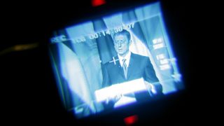 Blurry blue-toned broadcast screen showing a man in a suit delivering a speech, set against a dark background.