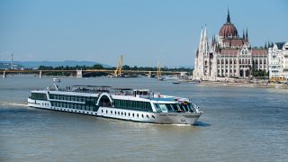 White passenger cruise ship on the Danube near Budapest's Parliament Building with a bridge and riverfront in the background.
