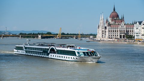 White passenger cruise ship on the Danube near Budapest's Parliament Building with a bridge and riverfront in the background.