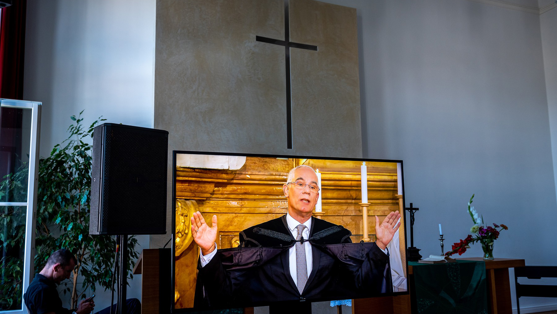 A preacher on a video screen delivering a sermon, raised hands, in a church setting with a cross backdrop and candles nearby.