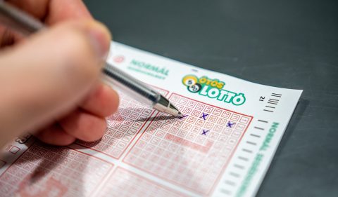 Close-up of a hand scratching a red-bordered lottery ticket; the LOTTO logo is visible near the top edge.
