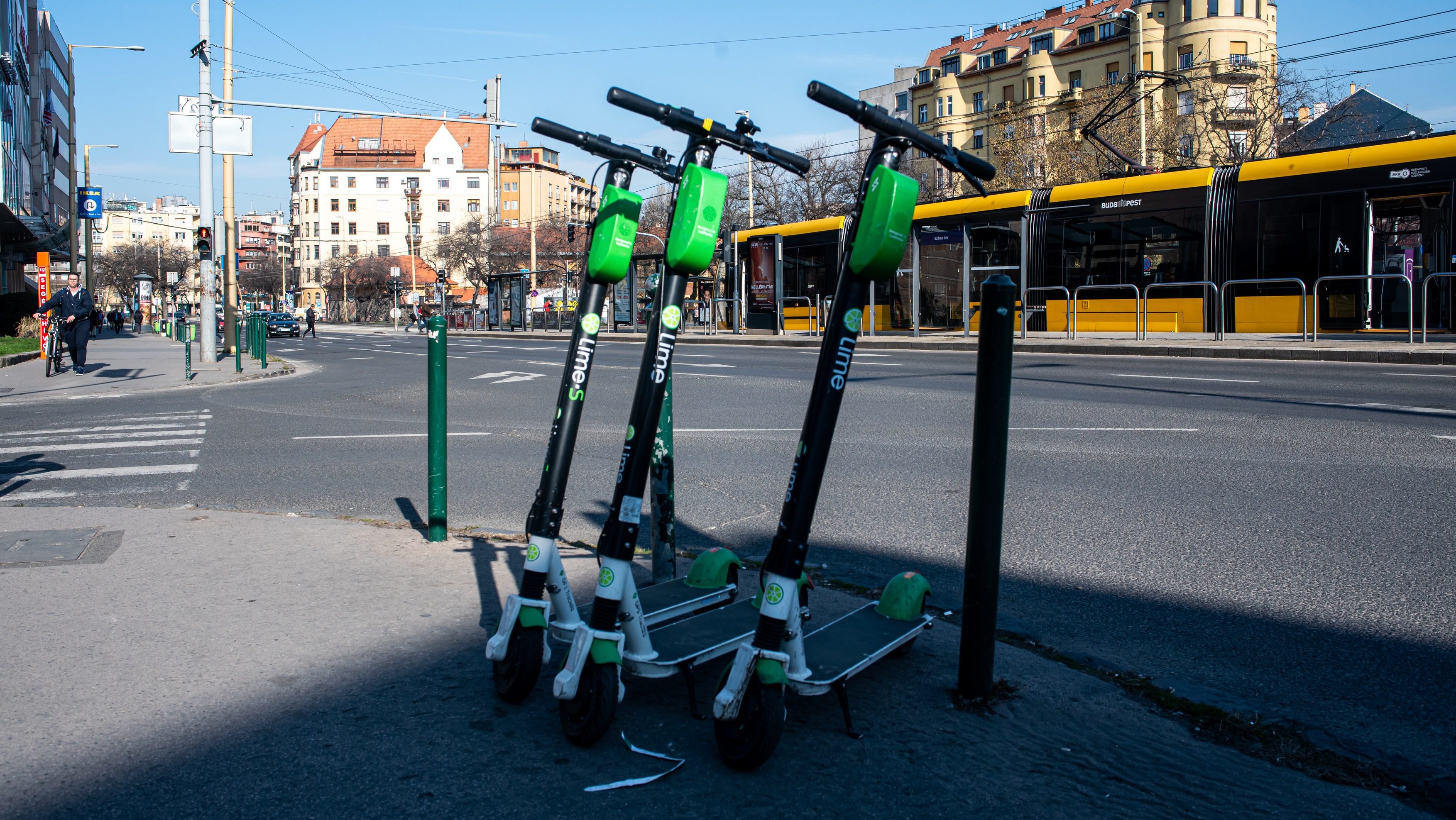 Green Lime e-scooters leaning together on a city street foreground with a yellow tram passing behind them.
