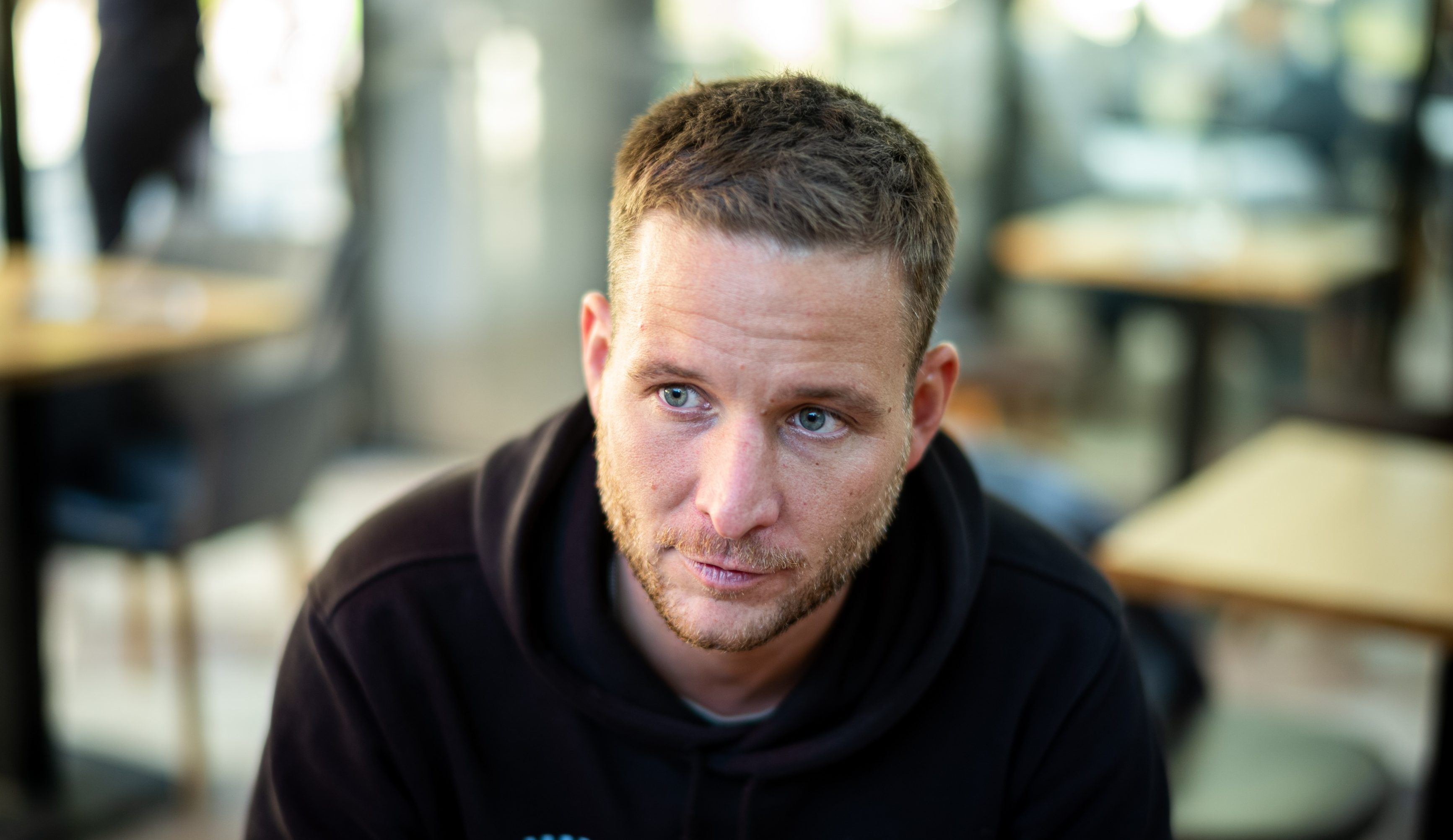 Close-up of a man with short brown hair and blue eyes wearing a black hoodie, looking to the left in a blurred indoor setting.