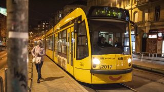 Yellow Budapest tram at a nighttime stop, with a woman in a pink coat waiting on the platform.